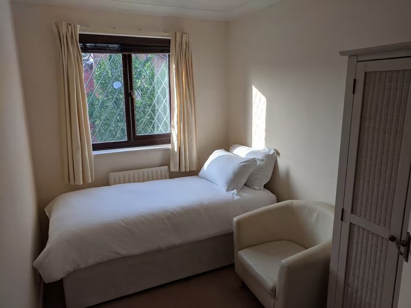 Bright bedroom with single bed, cream bedding, armchair, window with curtains