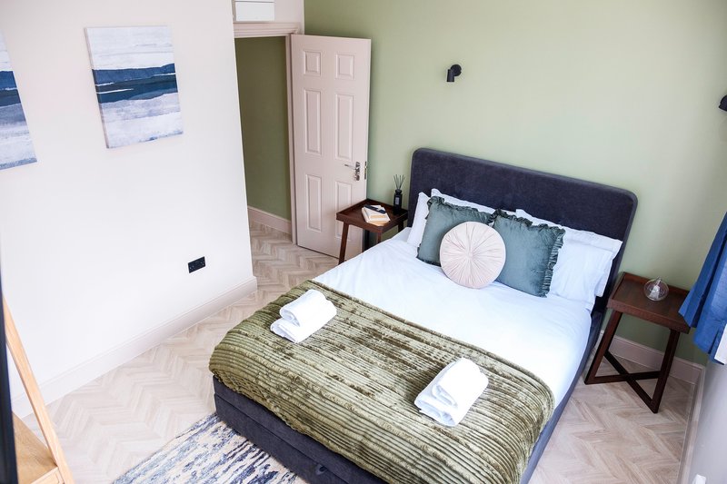 Bedroom with double bed, olive bedspread, white pillows, bedside tables, and coastal wall art