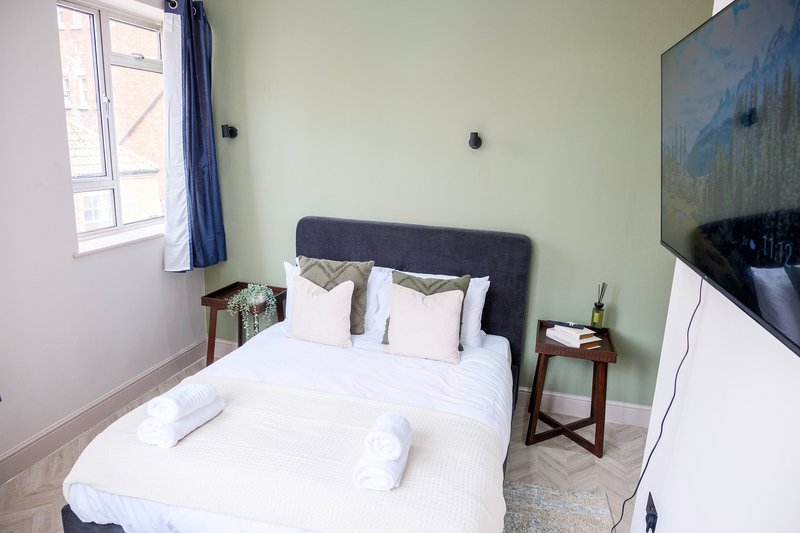 Bedroom with double bed, dark headboard, white bedding, wall-mounted TV and window with blue curtains