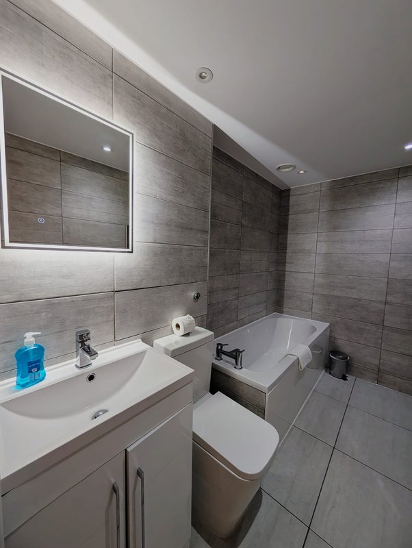 Modern bathroom with grey tile walls, white vanity sink, toilet, bathtub and illuminated mirror