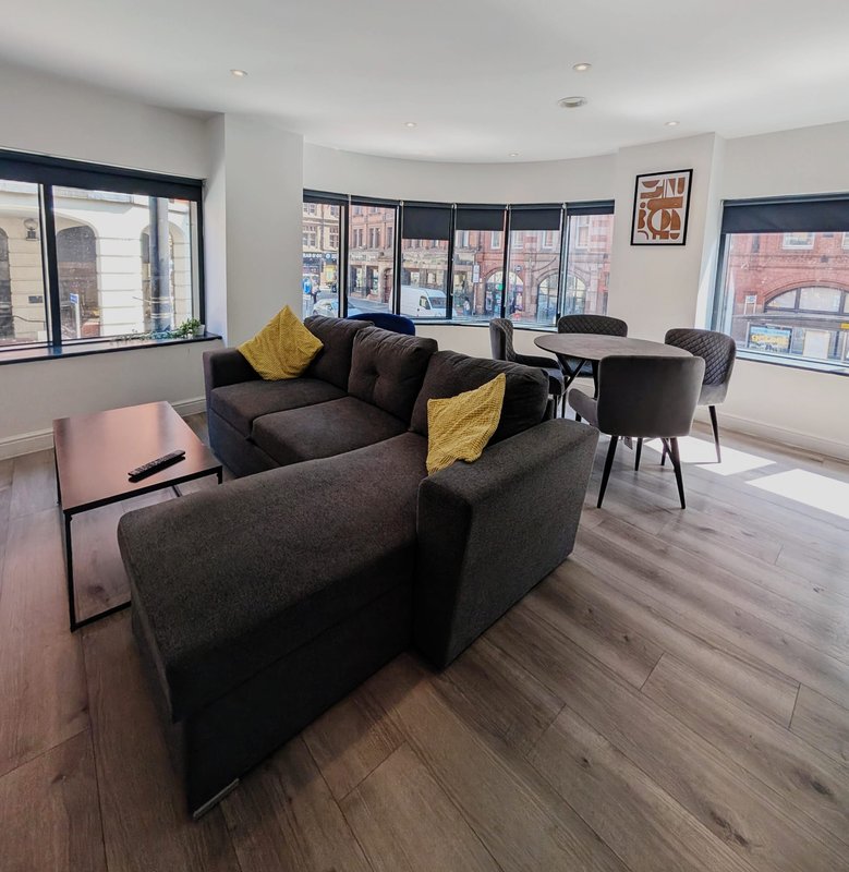Modern living room with dark sofa, yellow cushions, dining table, and city views from windows