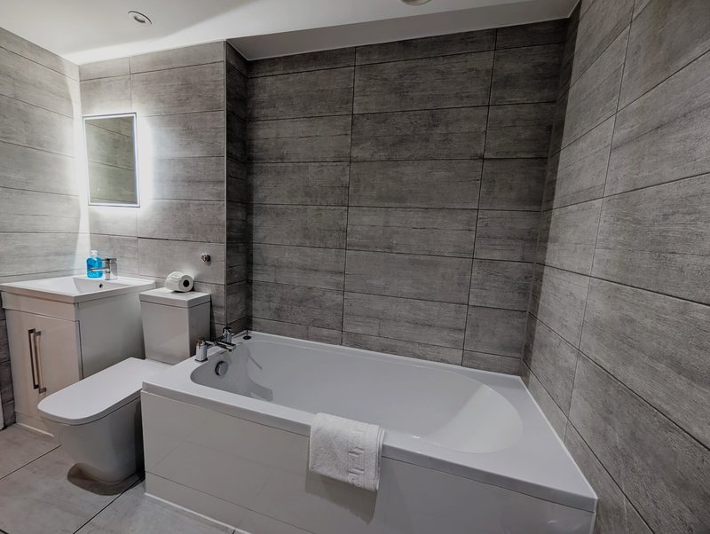 Modern bathroom with white bathtub, gray tile walls, toilet, sink and mirror