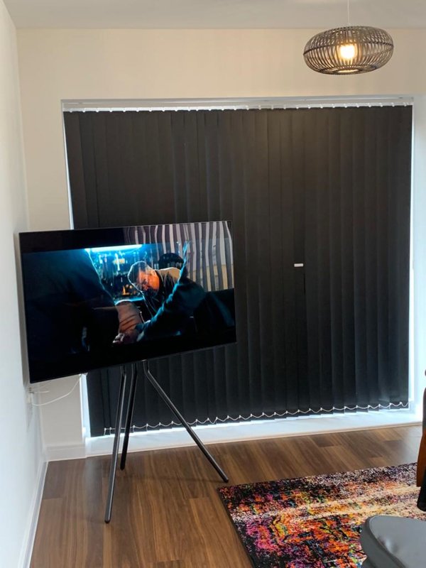 Living room with wall-mounted TV on tripod stand, wooden flooring, vertical blinds and ceiling lamp