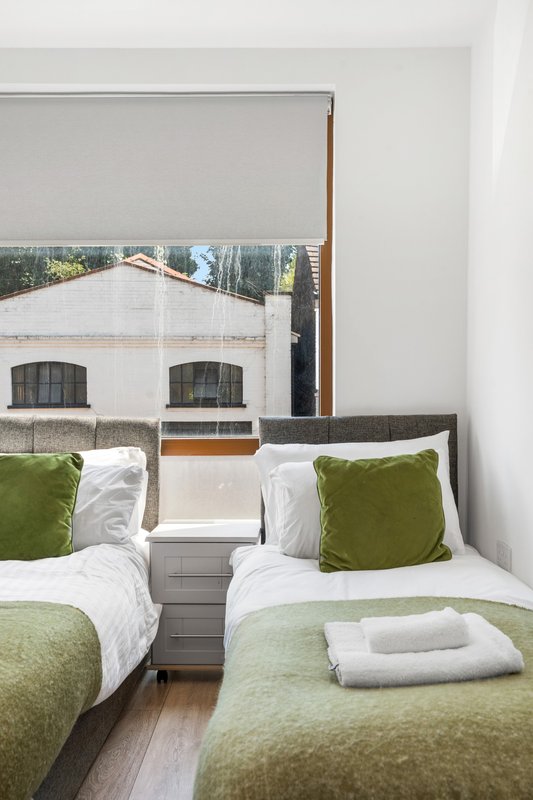 Modern twin bedroom with green cushions, white bedding, and window view of residential building