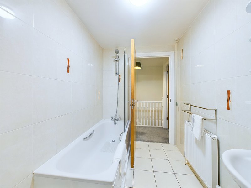 Narrow bathroom with white tiles, bathtub with shower, wooden accents and towel rail