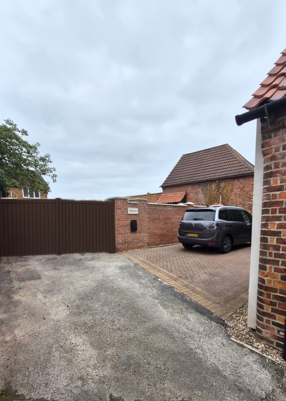 Brick driveway with parked car, brown gate, and residential buildings under cloudy sky