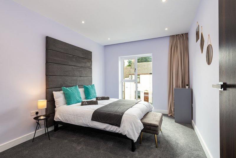 Modern bedroom with double bed, turquoise cushions, dark headboard, and glass door to garden