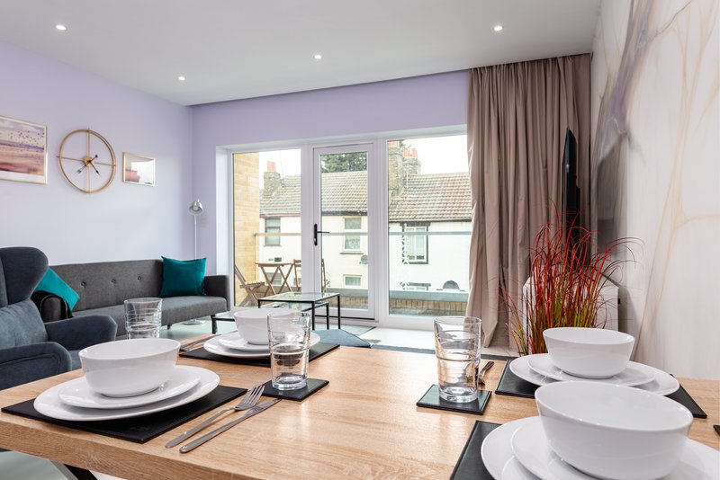 Open-plan living and dining area with lavender walls, wooden table set for dining, and grey sofa with patio doors