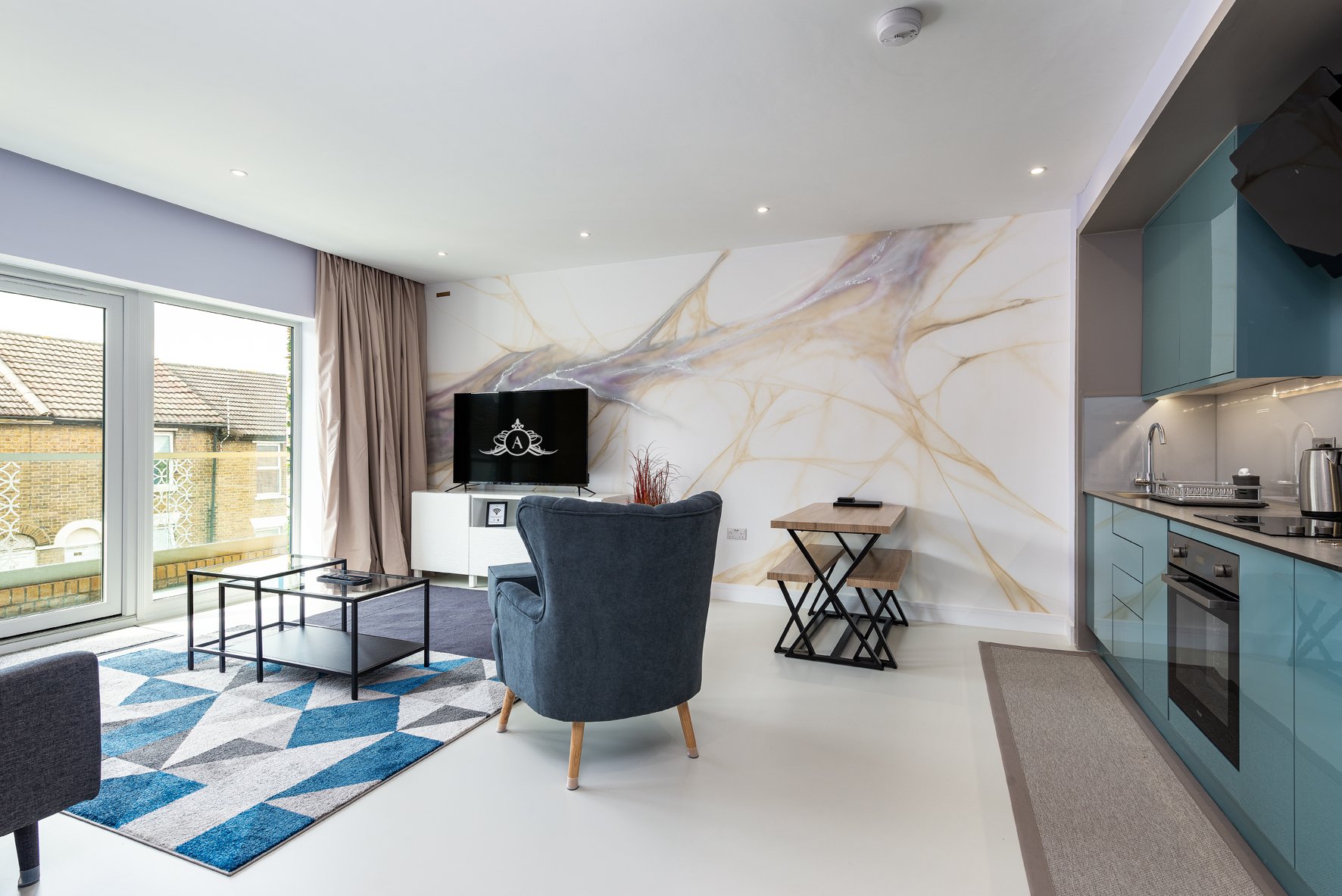 Open-plan living space with teal kitchen, marble accent wall, dark armchair and blue geometric rug