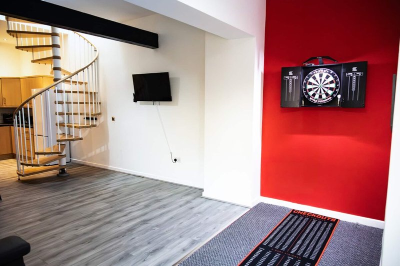 Open-plan living space with spiral staircase, TV mounted on wall, dartboard, and red accent wall