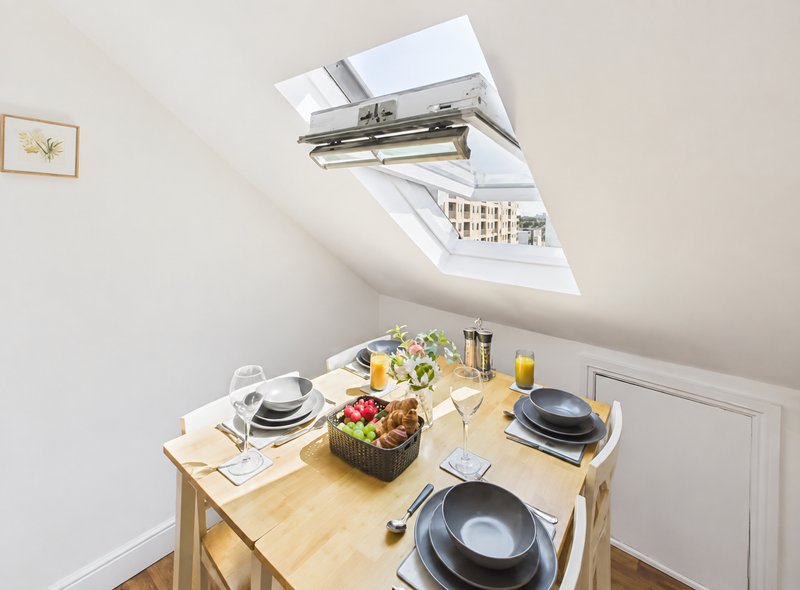 Bright attic dining area with wooden table, place settings, skylight window and fresh flowers