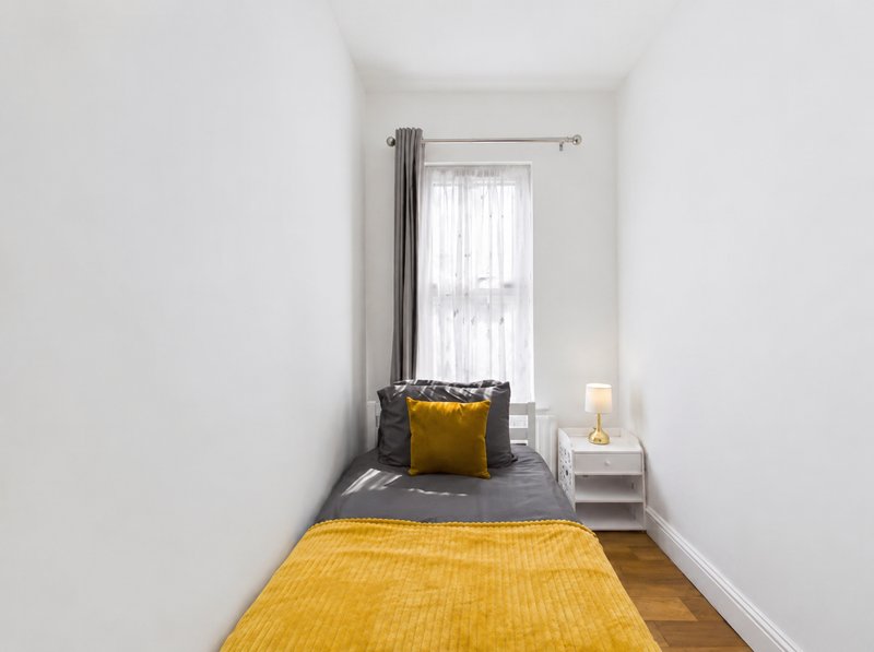 Narrow bedroom with single bed, yellow bedding, gray headboard, window with curtains, nightstand lamp