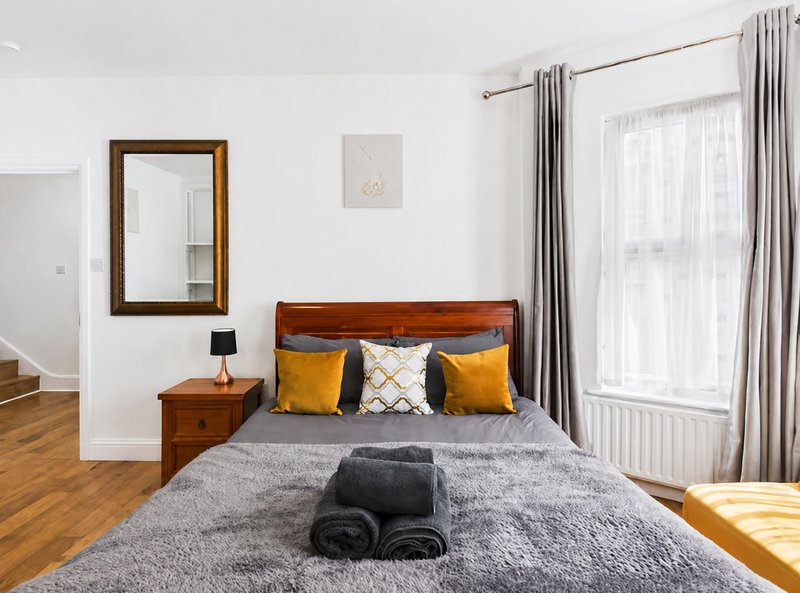 Modern bedroom with wooden bed frame, grey bedding, yellow cushions, mirror and window with curtains