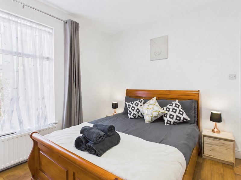 Bright bedroom with wooden bed frame, grey bedding, patterned cushions and large window with curtains
