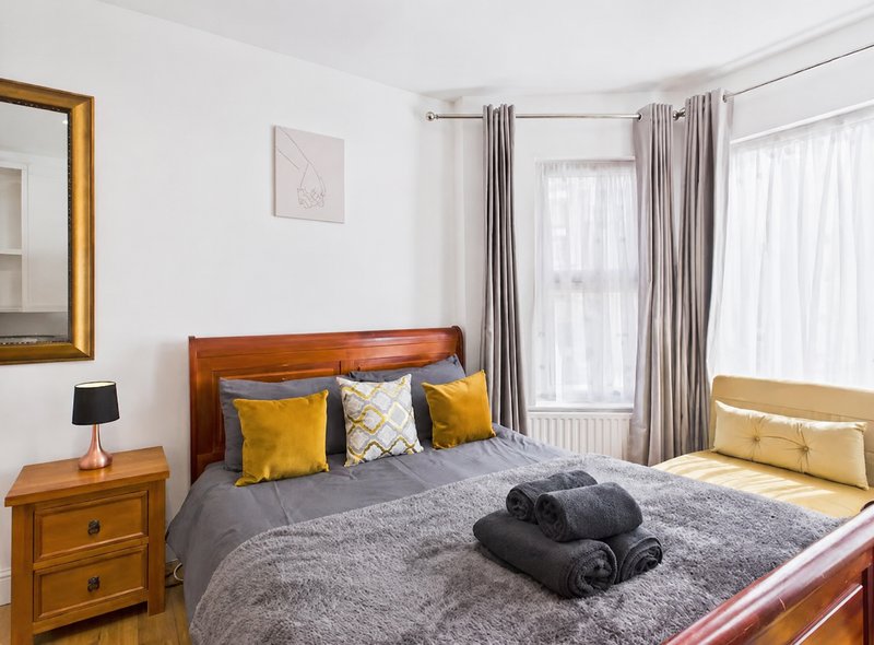 Bright bedroom with wooden bed, yellow and grey bedding, wooden nightstand, and floor-to-ceiling windows