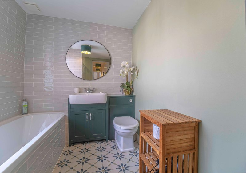 Bathroom with pink subway tiles, dark vanity, round mirror, bathtub and wooden storage shelf