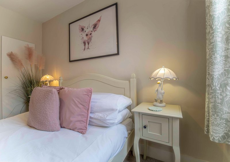 Bedroom with single bed, pink cushions, white bedside table, lamp and framed artwork
