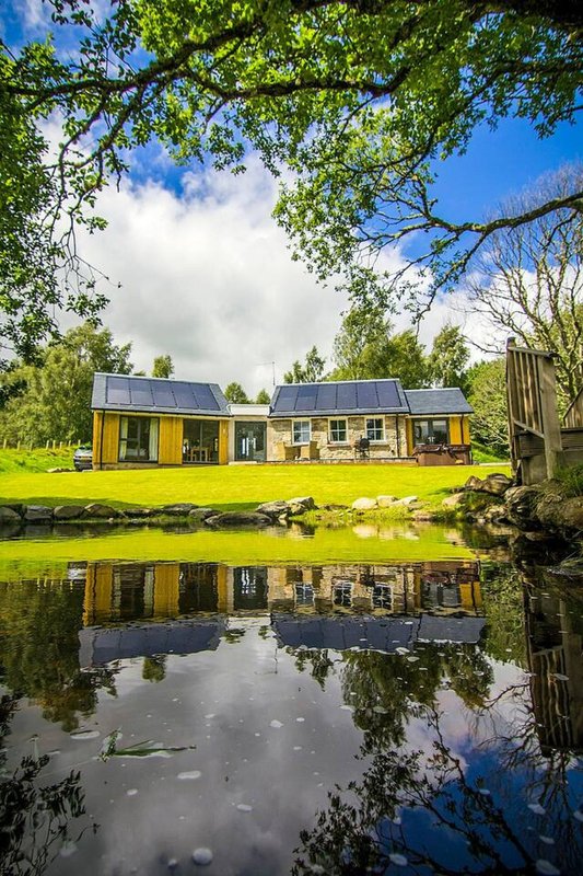 Modern yellow property with solar panels reflected in pond, surrounded by trees and green lawn