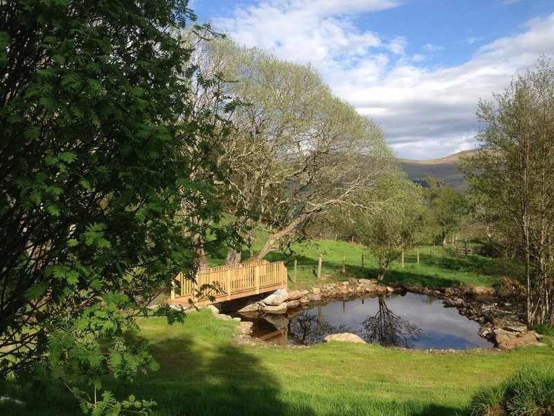 Rural garden with wooden bridge over pond, green lawn, trees and distant hills