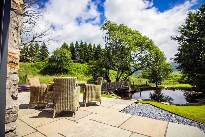 Patio seating area with wicker chairs overlooking pond, garden and tree-lined landscape