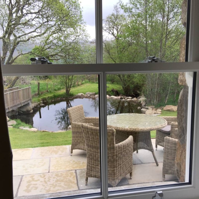 View through large windows to garden with patio furniture, pond, and surrounding trees