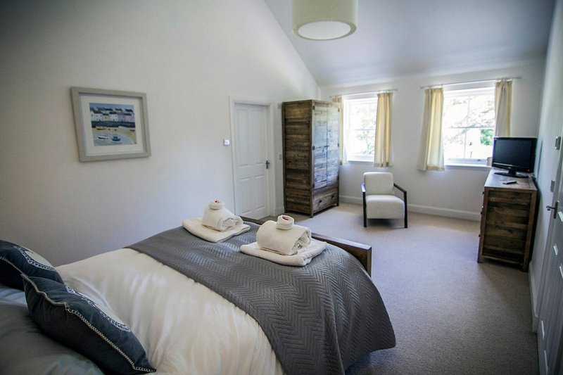 Spacious bedroom with double bed, white pillows, gray bedding, wooden chest of drawers, and seating area