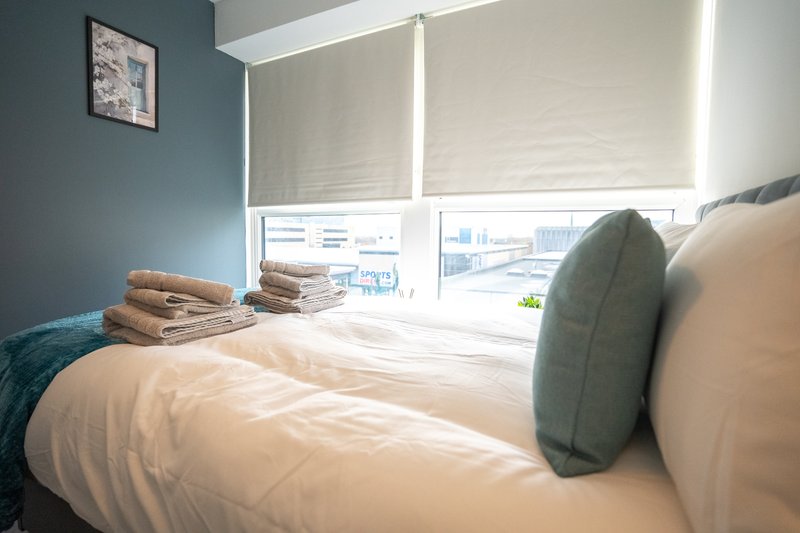 Bright bedroom with double bed, green pillows, rolled towels, and city view windows