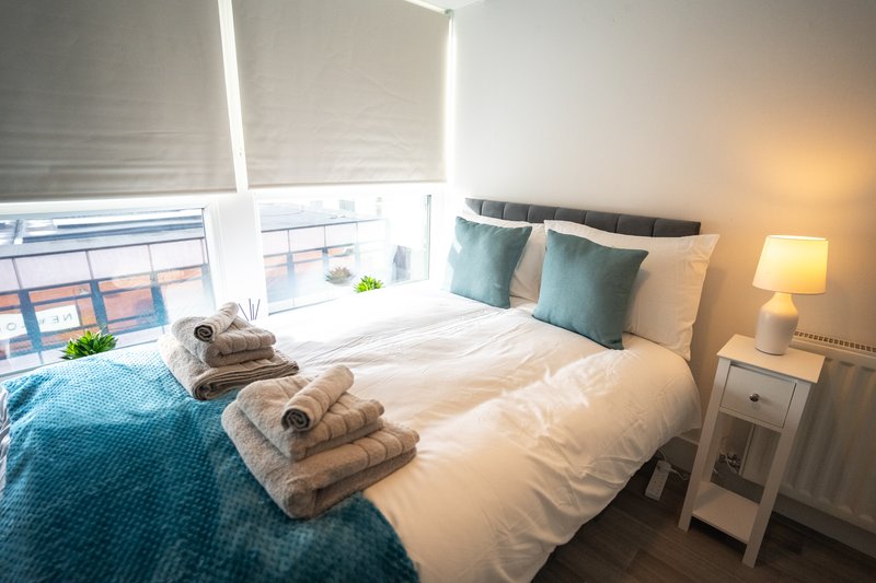 Bright bedroom with double bed, rolled towels, teal bedding, cushions, and window with blinds