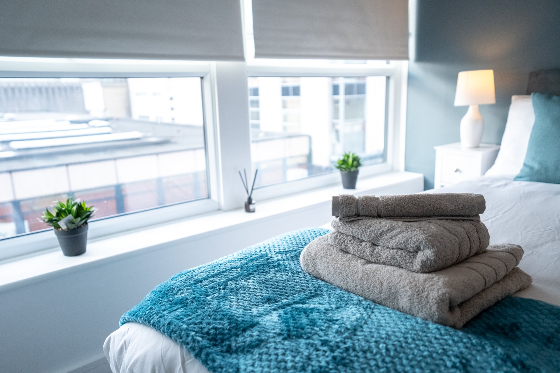 Bright bedroom with folded towels on bed, blue throw, large windows and potted plants