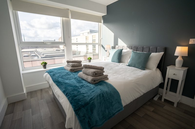 Modern bedroom with double bed, teal throw, stacked towels, dark accent wall and large window.