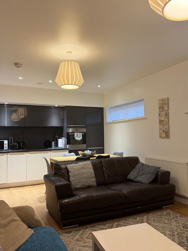 Open-plan living room with black leather sofa, kitchen area, pendant lighting and neutral decor