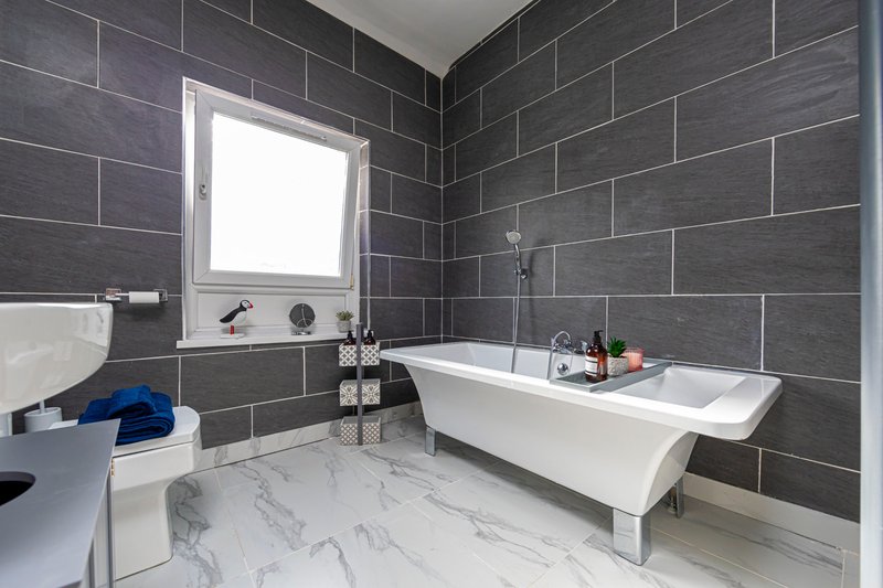 Modern bathroom with dark gray tile walls, white freestanding tub, toilet, and marble floor