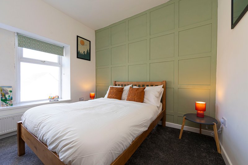 Bedroom with double bed, green panelled feature wall, wooden bedframe, and bedside table with lamp