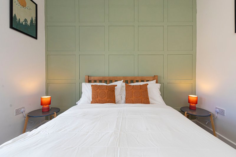 Bedroom with double bed, white bedding, orange cushions, green panelled headboard and bedside lamps