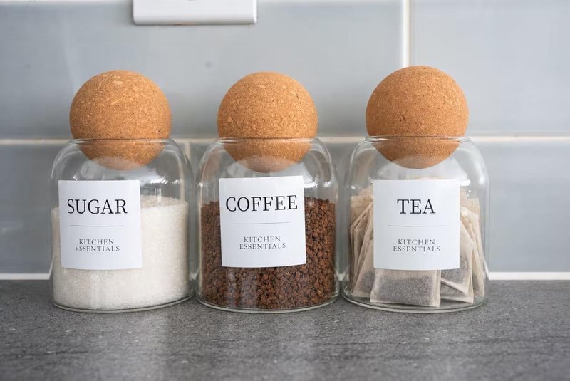 Three glass storage jars with cork lids containing sugar, coffee, and tea on kitchen counter
