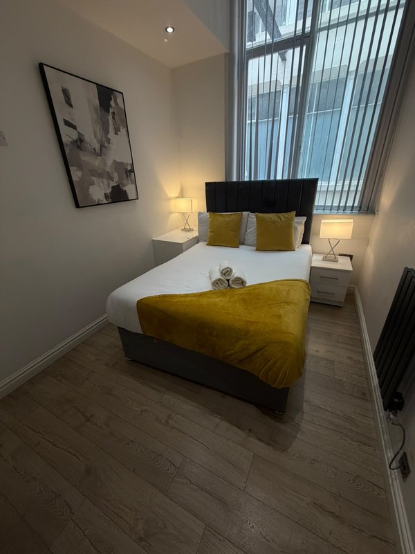 Modern bedroom with double bed, black headboard, yellow bedding, white pillows, bedside lamps, wall art and vertical blinds