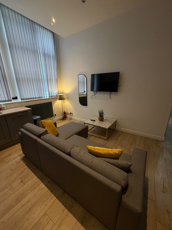 Modern living room with gray sofa, wall-mounted TV, yellow accent cushions, and natural light from vertical blinds