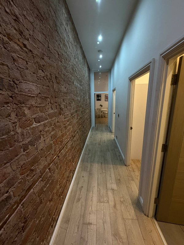Long narrow hallway with exposed brick wall, wooden floor, recessed ceiling lights, and closed doors