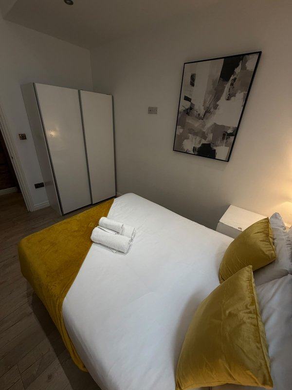 Bedroom with double bed, mustard-yellow velvet headboard, white bedding, wardrobe, and wall art
