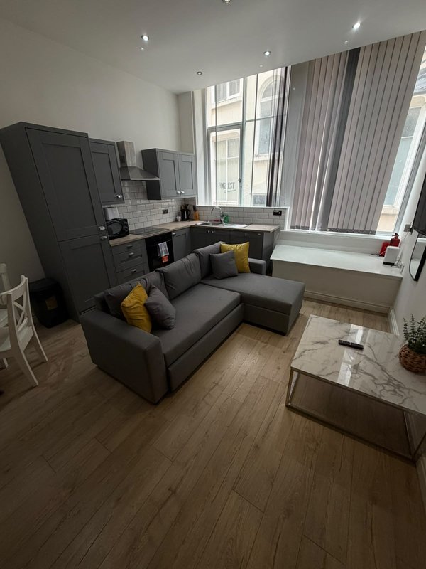 Modern open-plan living area with dark sofa, kitchen units, and large windows with curtains