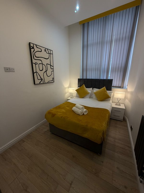 Compact bedroom with mustard yellow bed, black headboard, white bedside lamps and abstract wall art