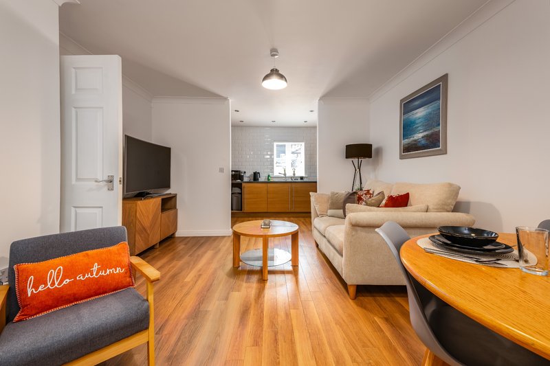 Open-plan living room with cream sofa, wooden furniture, TV, and kitchen visible in background