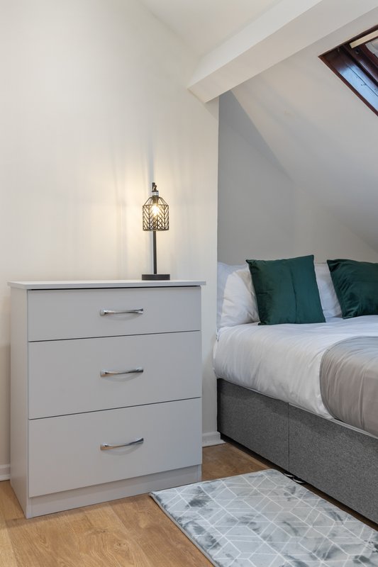 Bedroom with white chest of drawers, decorative lamp, bed with green cushions and white bedding