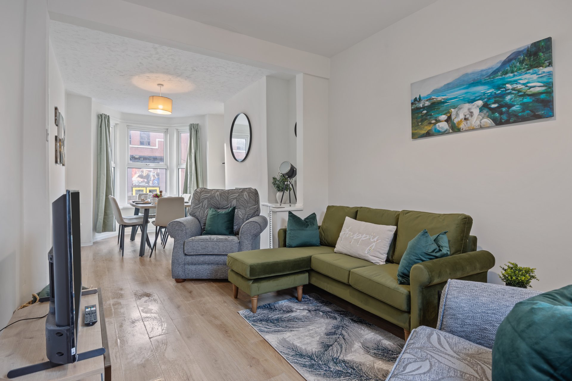 Open-plan living room with green sofa, grey armchair, wooden flooring and coastal artwork