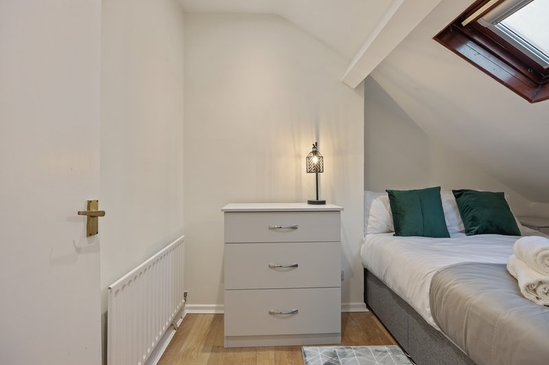 Small attic bedroom with white chest of drawers, double bed with green pillows, and skylight window