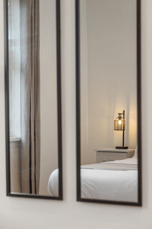 Bedroom reflected in two mirrored panels, showing bed with white linens and bedside lamp