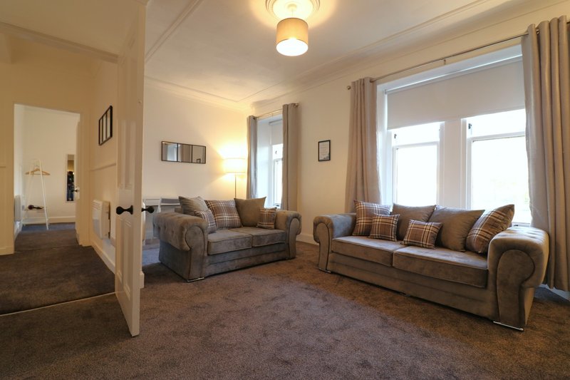 Spacious living room with two grey sofas, brown carpet, large windows with curtains, and ceiling lights