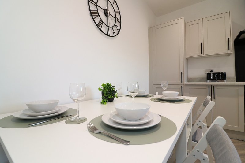 Modern dining area with white table, place settings, decorative clock and kitchen cabinets