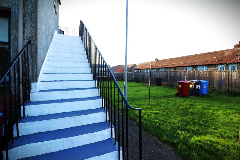 Exterior staircase with blue and white steps leading to grassy courtyard with bins and terraced houses