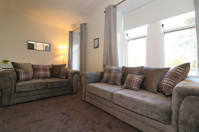 Bright living room with two grey sofas, plaid cushions, lamp, and large windows